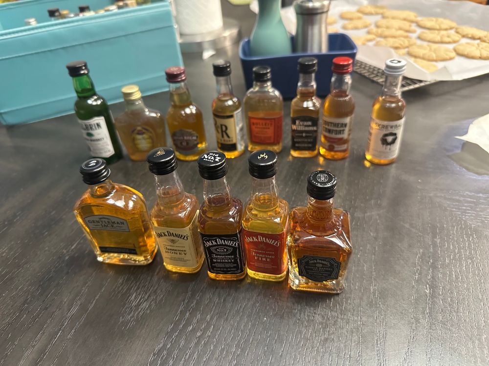 A dark gray table with 13 taster size bottles of whiskey on it in two rows. The front row is five different Jack Daniel’s whiskies, and the back row is a smattering of other brands and types of whiskey. Two of the bottles in the back row are bourbon, which tastes very bitter to me because of some compound in the barrels, so I didn’t taste those. There is a light turquoise box in the back left with some other spirits taster bottle caps peeking out from the top. There is also a roll of paper towels, a salt and pepper shaker, and a cooling rack covered in parchment paper and cookies all in the background as well. Most of the photo is out of focus.