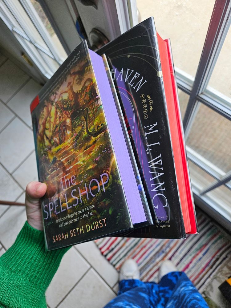 A hand holds The Spellshop by Sarah Beth Durst with a purple sprayed edge, and Blood Over Bright Haven with a red sprayed edge. 