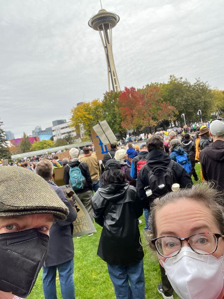 Selfie of 2 white people in N95s in front of a peaceful protest at the Space Needle.