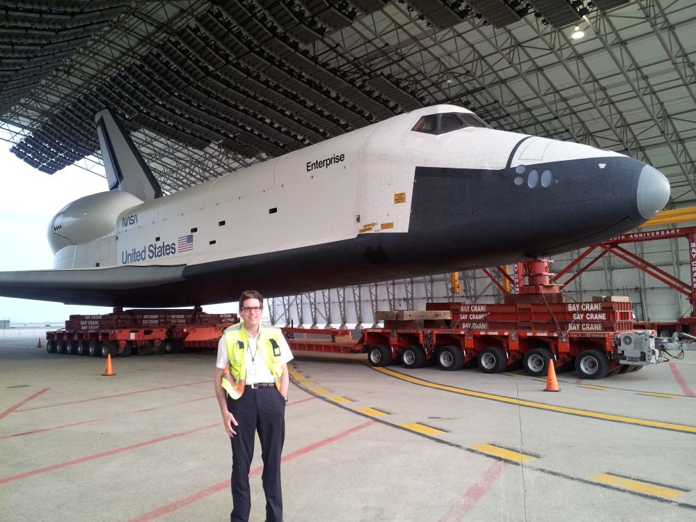 A man in a high visibility vest standing in front of the Space Shuttle Enterprise loaded on a truck.