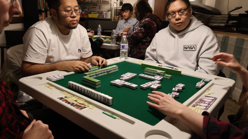 A game of riichi mahjong. Two tables of 4 people each are playing simultaneously. Someone has just won and is showing her hand. The 3 other players at her table are examining it thoughtfully.