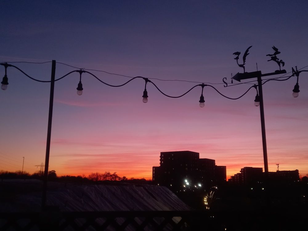 A dark blue, to pink, to orange sunset. With a few stripes of clouds. And some buildings, lights and a weather vane are silhouetted against the vibrant sky.