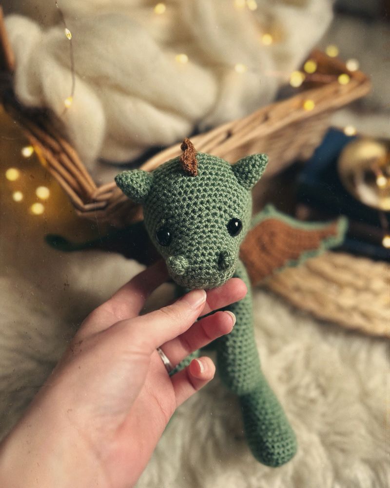My hand holding the head up of a green and brown crochet dragon. 