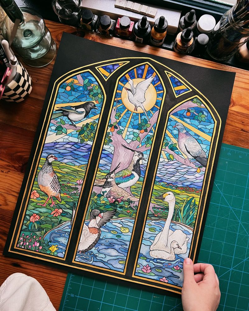 Picture of Julie’s large watercolor piece “St. Patrick’s Green” inspired by stained glass and featuring seven bird species found in Ireland. 

The painting is a landscape featuring a duck and swan in a pond, a pheasant and pair of lapwings in the field, a magpie and woodpigeon in a tree, and arctic tern flying in the sky. In the background are mountains and a bright sun. 