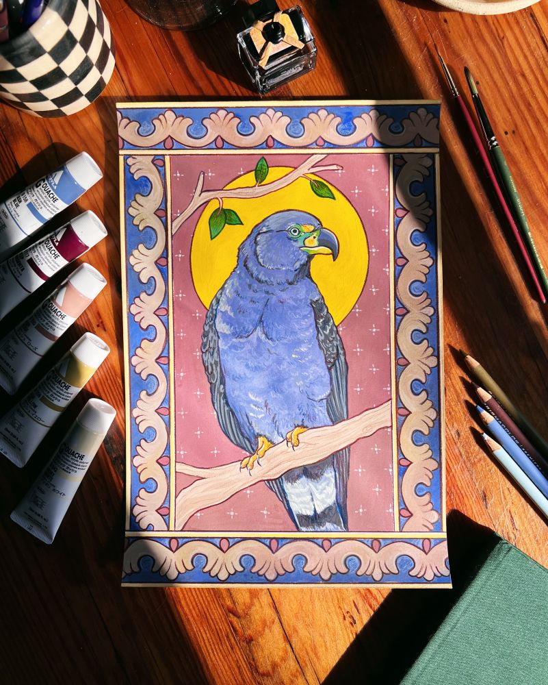 A flat-lay photo of a painting on a desk, surrounded by artist materials like Holbein Gouache and Prismacolor colored pencils. The painting is of a hook-billed kite against a background of patterns inspired by medieval illuminated manuscripts 
