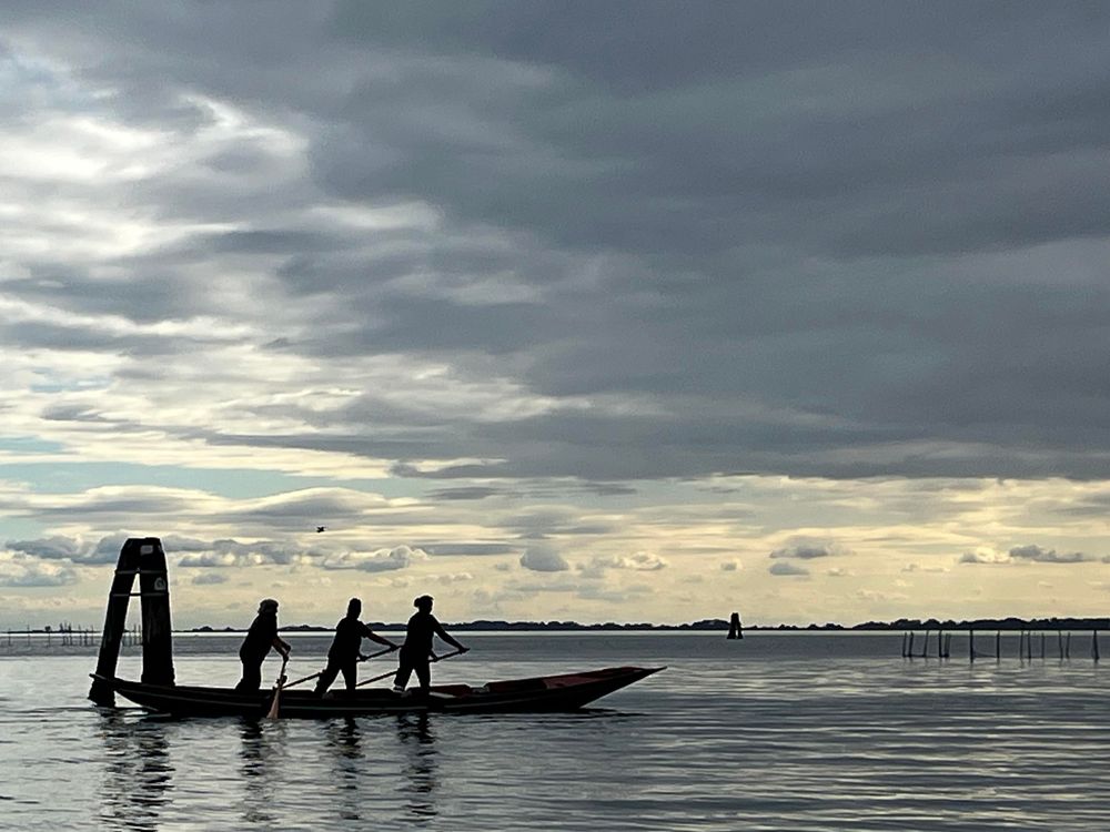 The Venetian lagoon, silver water under a sliver cloudy sky, lit by a weak sun into a strip of mother-of-pearl above distant tree-lined island in the far distance along the horizon line. In the foreground on the right of the frame, in dark black silhouette, a three-legged wooden navigation post, and a low black racing gondola, being rowed by three standing figures, their legs braced apart one in front of the other, their arms stretched out holding the long oars at an angle.