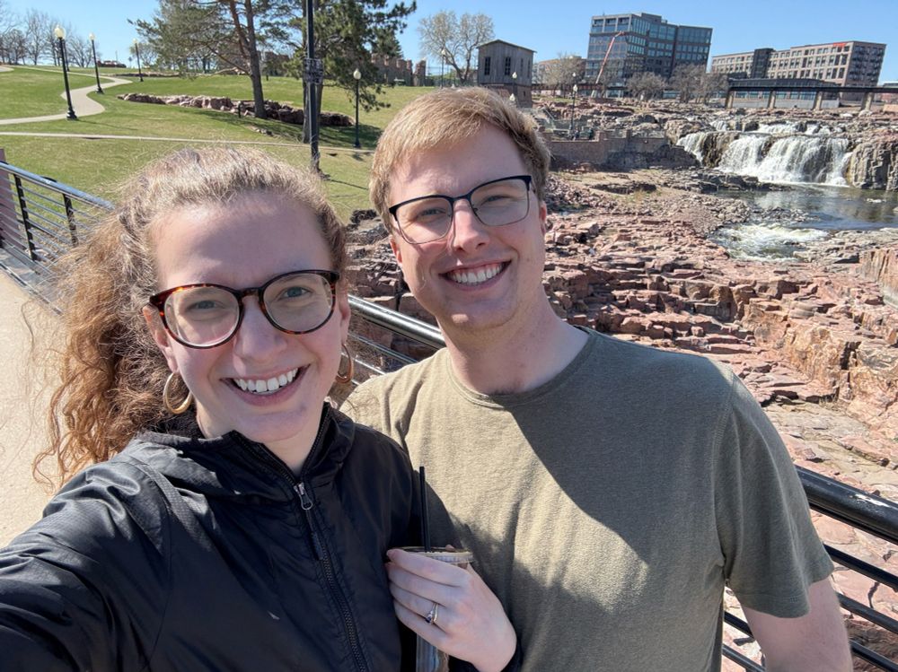 Woman in black jacket and glasses and man in gray tee shirt and glasses standing in front of a waterfall with a cityscape in the background. 