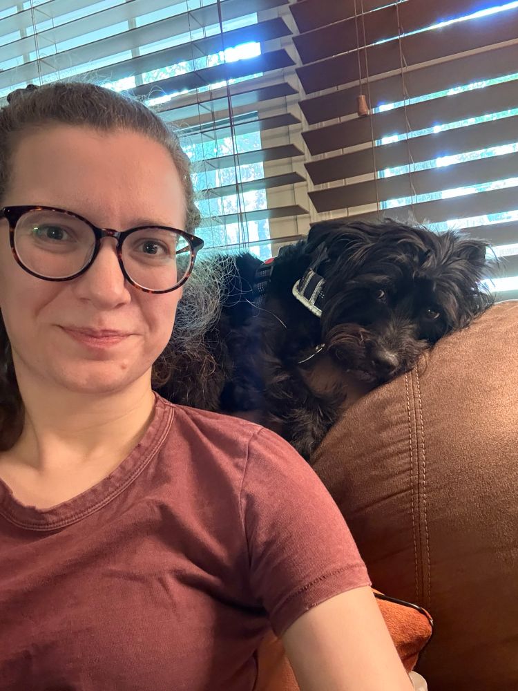 Woman sitting on a brown sofa with a little black scruffy dog laying on top of the sofa giving the woman side eye. 