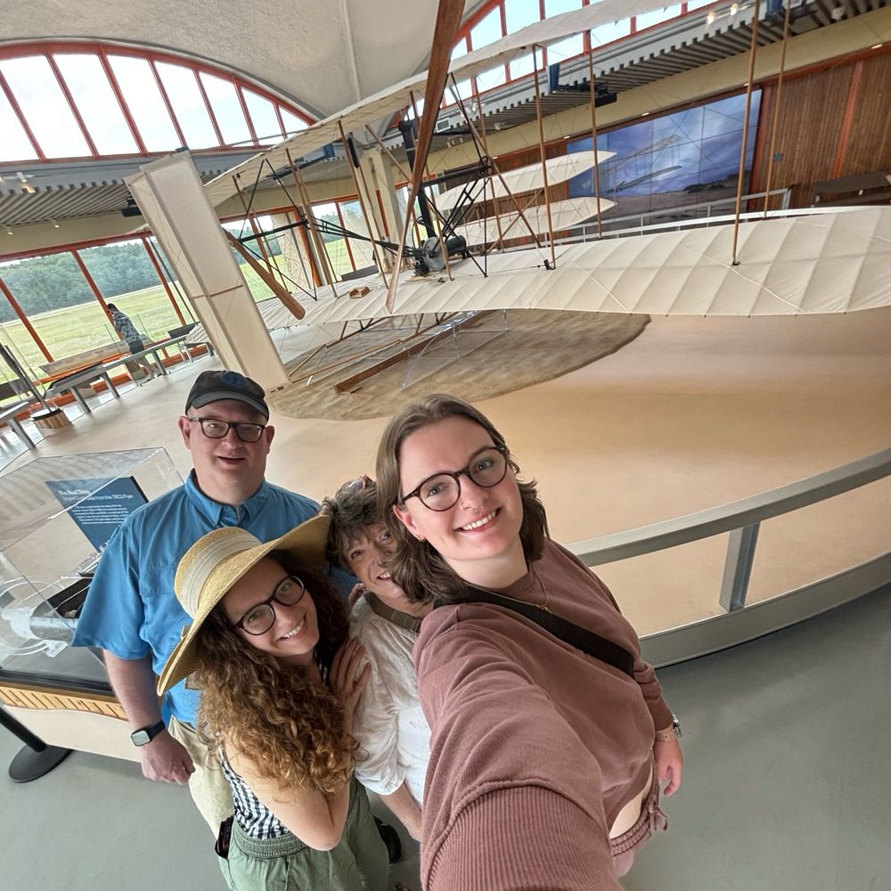 A man and three woman stand in front of a life-size museum display of an early airplane 