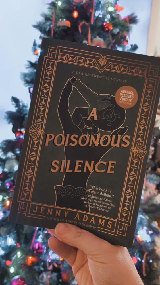 Green and gold book (A POISONOUS SILENCE by Jenny Adams) held up in front of a decorated christmas tree.