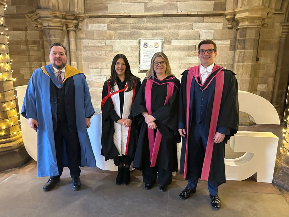 Four staff from Information Studies. We are all wearing academic gowns. From left: David wearing a blue gown with a yellow and blue hood; Kat wearing a black gown with wide white trim with red details and a black and red hood, then Adele and Ian wearing a black gown with pink trim and a pink and black hood (the Glasgow PhD gown).