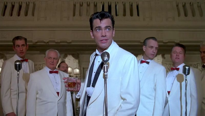 Peter Gallagher starting to sing Memories Are Made Of This while holding a drink and wearing a white dinner jacket, as Vic Tenetta in THE HUDSUCKER PROXY