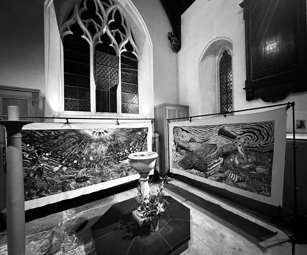 Two large modern artworks hang in an ancient church, with arched Gothic windows behind and a font in the foreground. The artworks are black-and-white prints on canvas, both celebrating the beauty and diversity of sealife along with the manmade threats - of plastic and other pollution in the first; various forms of seabed destruction in the second. Atmospheric black and white photo, taken by the vicar after a harrowing day and a midnight hang: peace reigns.
