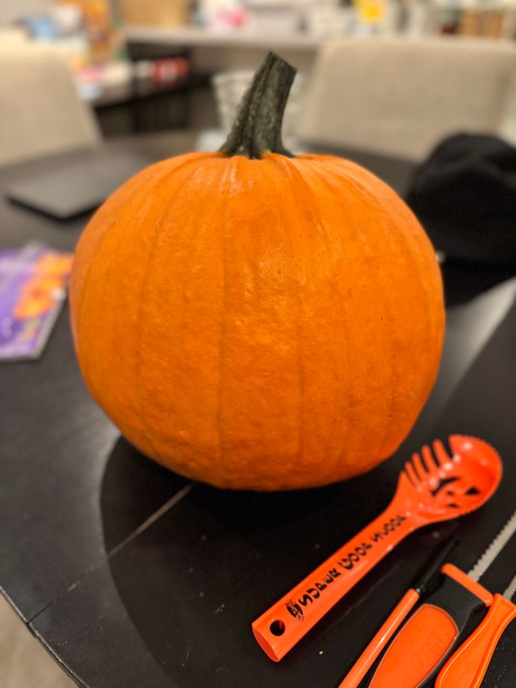 A remarkably round pumpkin sits atop a black table, carving tools lay to its right. 