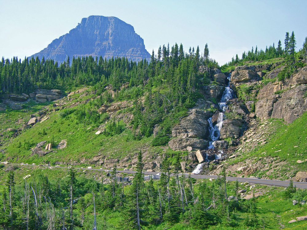 A paved highway runs across the image, below it is a lightly treed slope, across it there is a steeper slope with grasses leading up to evergreens on the left, on the right a waterfall tumbling through a rocky gorge then a steep rock slupe. Above, on the left is a mountain, steep and rocky. Overhead, the sky is blue.