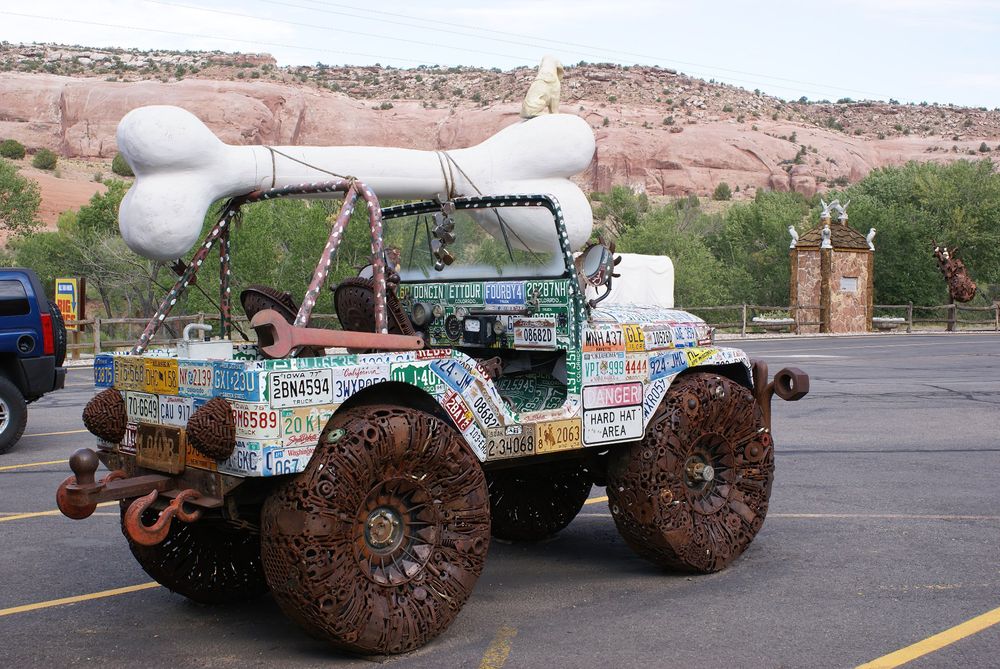 A scrap metal jeep sculpture. It is carrying a large white bone, a dog sits atop the bone