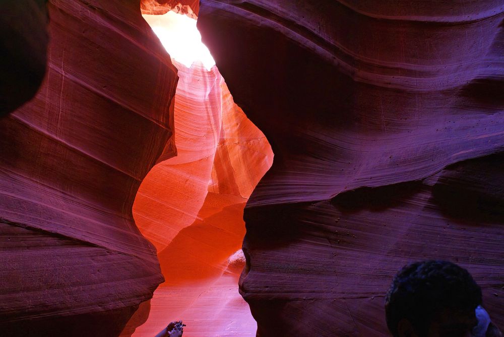 Looking through a narrow gap where both walls of the canyon appear a deep violet into l more brightly lit section which is glowing orange. On the lower right is a silhouetted head where someone crept  into the image