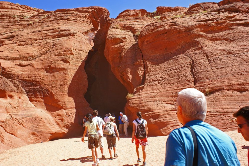 A group of people entering a narrow slit in a sandstone cliff. They are walking over near white sand. A small sliver of blue sky overhead.