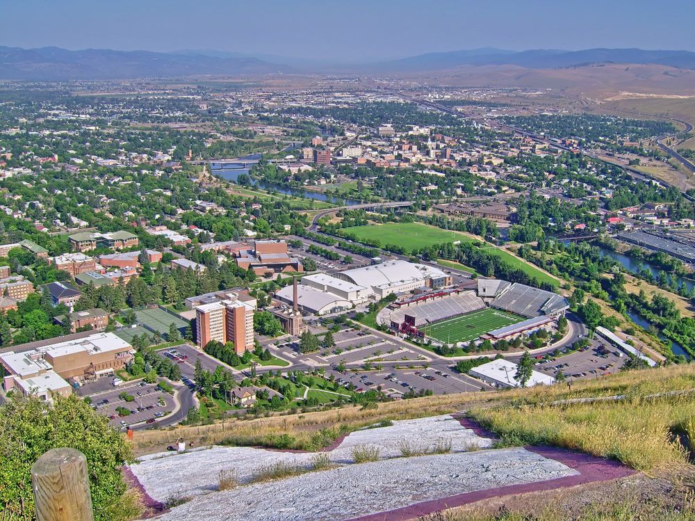 Scene overlooking a city with a river running through it. In the immediate foreground is half of a large concrete "M" on a downslope. Just at the foot of the M, a couple of people are sitting on the grass enjoying the view. At the bottom of the slope are university buildings including a tall residence building and sports stadium. In the distance, beyond the city, lad rises into surrounding hills / mountains. Overhead a narrow band of hazy blue sky. And there is no thought that an ignorant orange clown could possibly become US President.