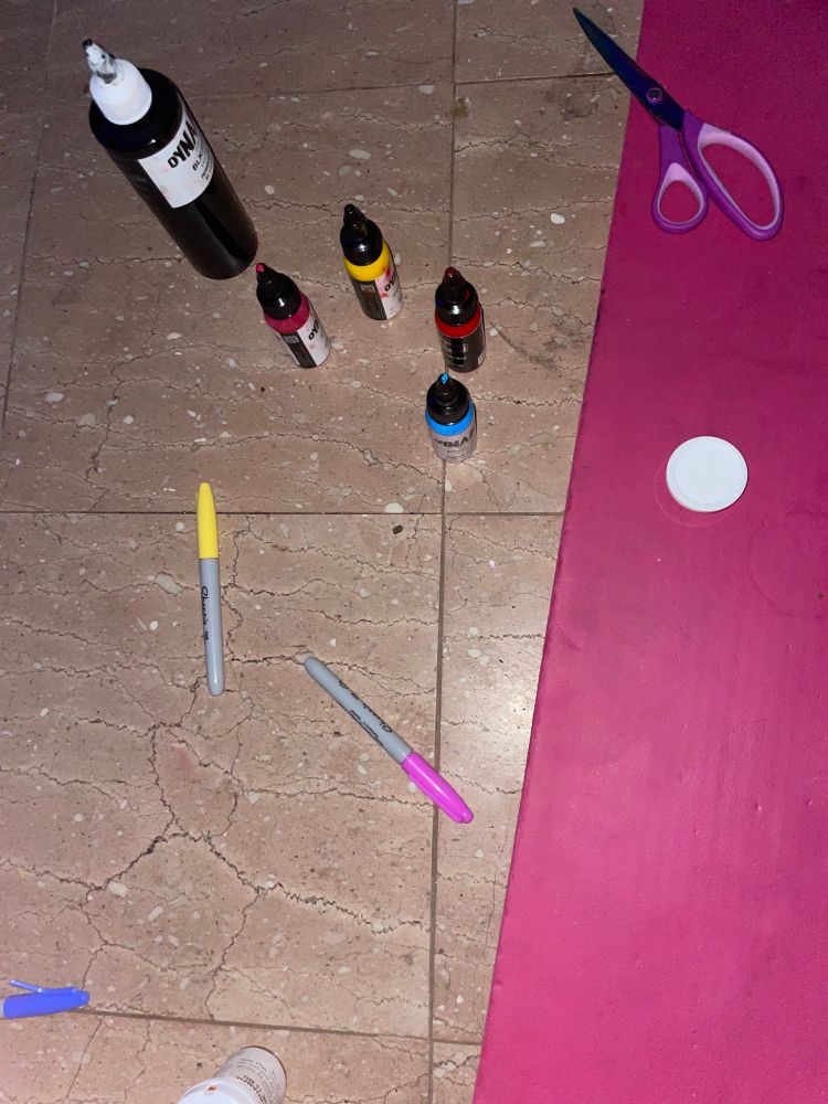 an assortment of brightly colored sharpies, bottles of bright tattoo ink, scissors, a bottle of prescription pills, and a pink yoga mat. the floor is dirty. 
