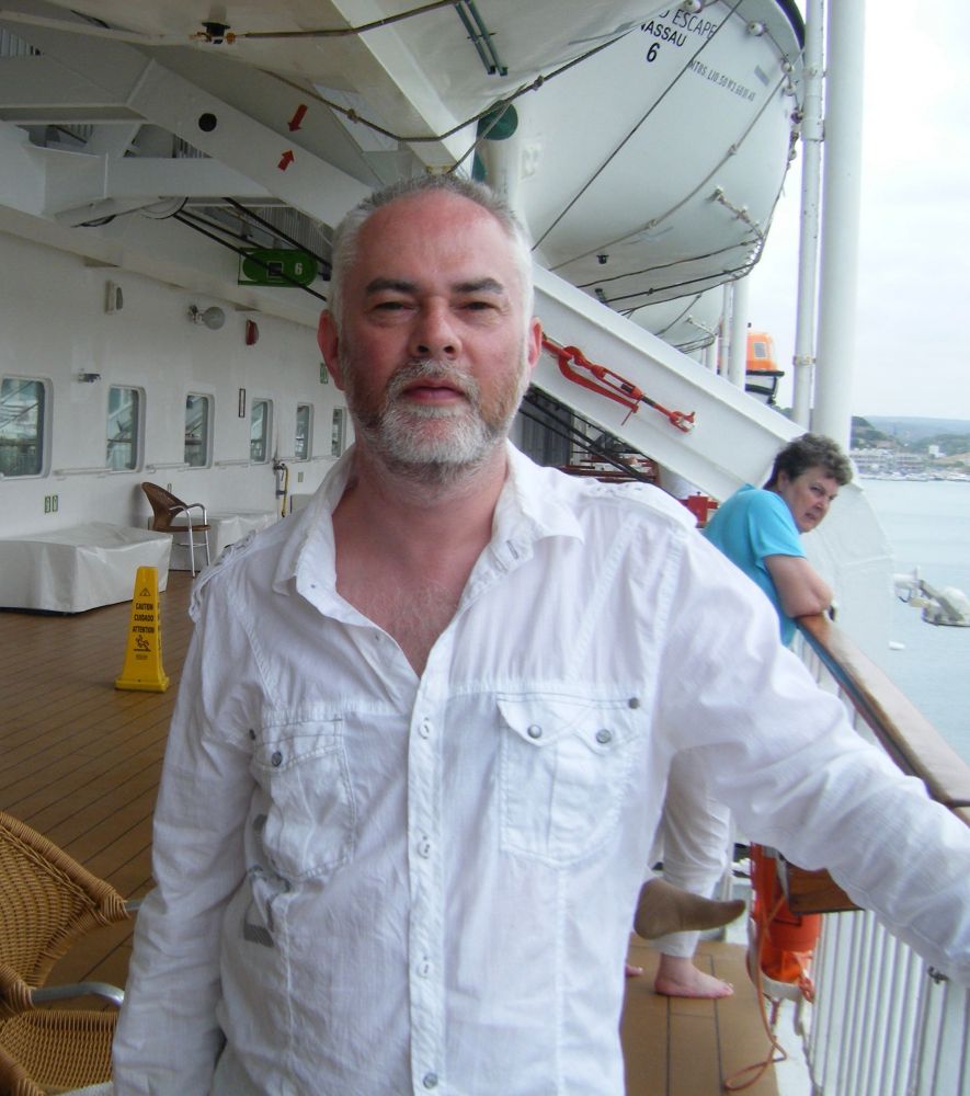 a bearded, balding man stood on a ship.  he is squinting because of the sunshine 