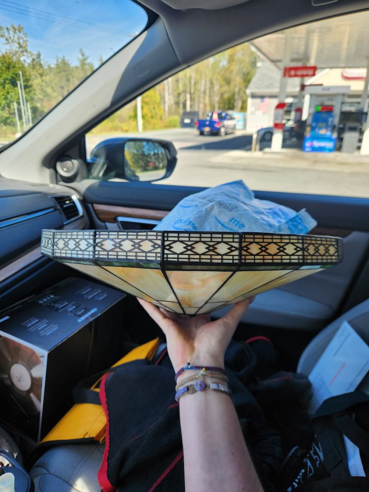 A leaded glass shade for a ceiling light being held aloft by a thin white human hand. The light is a shallow cone of creamy slag glass, with a thin green stripe at the edge and a diamond pattern border. The photo is in the shotgun seat of a car and out the window there's more blue sky and trees.