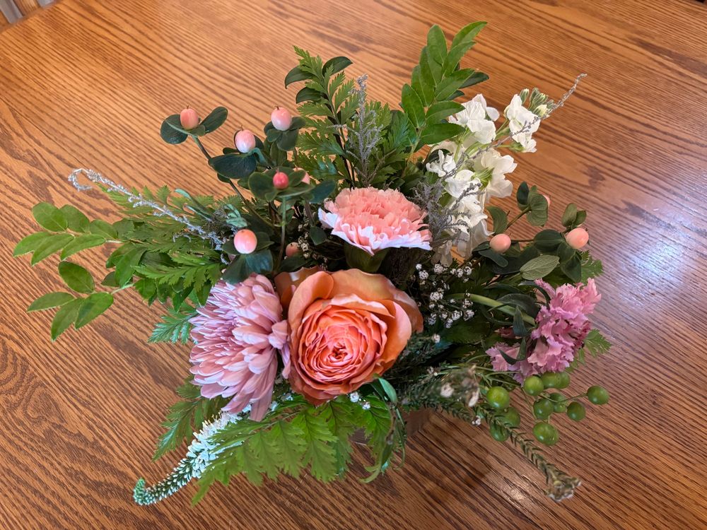 A fresh flower arrangement of greens and pink sitting on a brown table. 