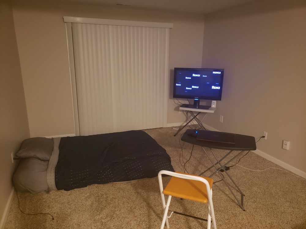 Photo of an apartment with a mattress on the floor, a folding chair next to a folding table and a tv on another folding table in the corner