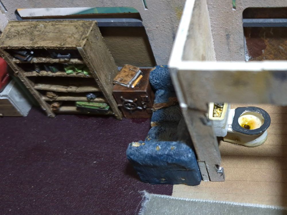 The inside of a 28mm residential trailer with a disgusting toilet, a ratty sofa, slightly rusted safe and a broken bookcase cluttered with tat
