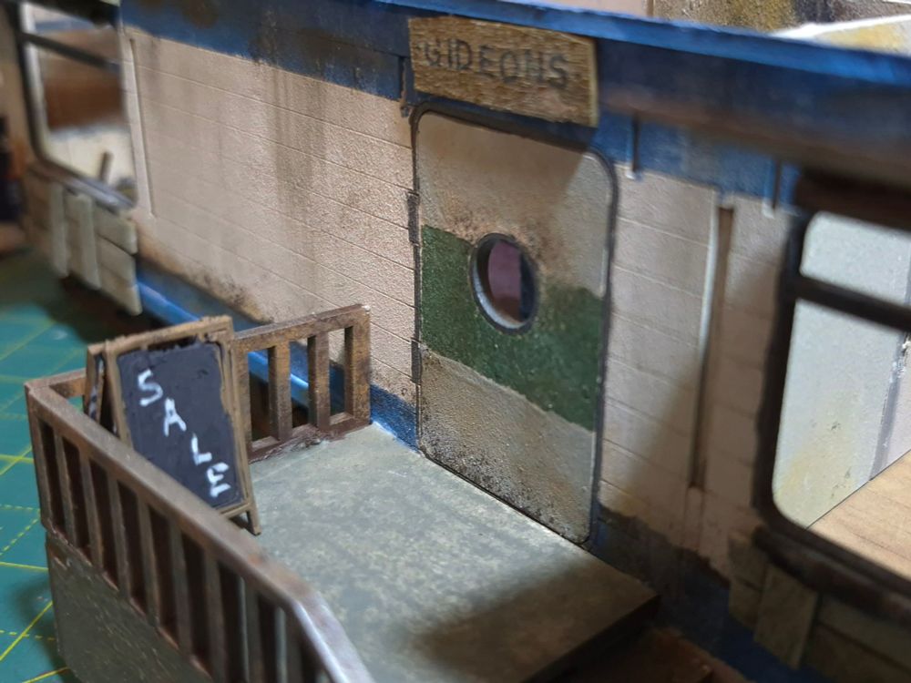The outside door of the residential trailer, a nameplate made from wood over the door reads "Gideons" and a sandwich board with "SALE" written on it stands outside 