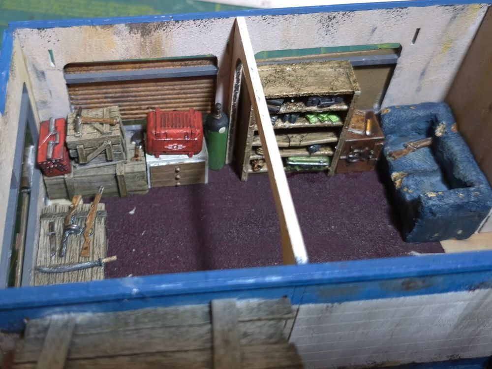The inside of the 28mm residential trailer there are piles of Crates of various sizes, a bookcase, a safe and a ratty sofa. Various items are laid out on top of the crates