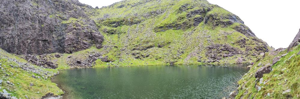 A deep green mountain tarn (small lake). The tarn is surrounded by dark rock on the back left and bright green grass on the other sides
