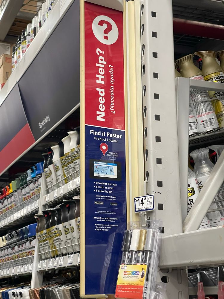 A vertical sign in a hardware store aisle displays a red and blue design with text in English and Spanish. The top portion reads “Need Help? ¿Necesita ayuda?” with a large question mark icon. The lower section promotes a product locator app, instructing customers to download the app, search for an item, and follow the pin to find products. Below the sign, a pack of DecoColor metallic paint markers is hanging on a hook. Spray paint cans are neatly arranged on shelves to the left.