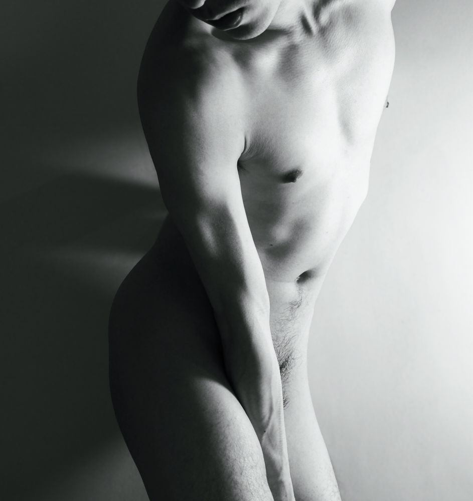 Bnw pic with high contrast and the light source from the right, casting rich shadows on the left side. The body is facing the light but twisted towards the opposite side. One arm is tucked between the thighs and the lower quarter of the face is visible too.