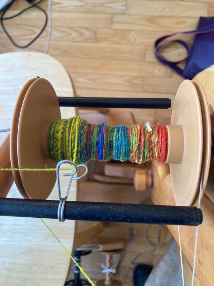 Spinning wheel in use. Close up of the bobbin with yarn on it. Multicoloured singles from a mixture of Merino and Shetland that I blended on my blending board. Yellow, red, blue, yellow green Merino, and hot pink (Shetland locks).  