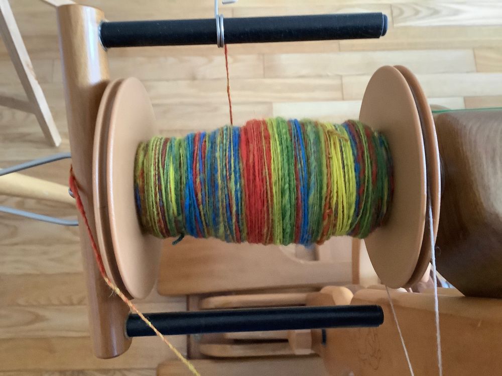 Spinning wheel bobbin on the wheel, with a multicoloured yarn of yellow, red, blue, green and a shot of hot pink locks on it.  