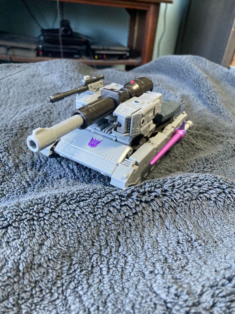 A photo of Studio Series 86 Leader Class Megatron in his Alt-mode, which takes the form of a light-grey tank. The tank’s turret is comprised of his fusion cannon, with the barrel formed from the one resting on his back on robot mode. next to it is a pistol resembling Megatron’s original Walther P-38 alt-mode, and the laser blade he uses against Prime in the 86 movie is slotted on the side of his treads
