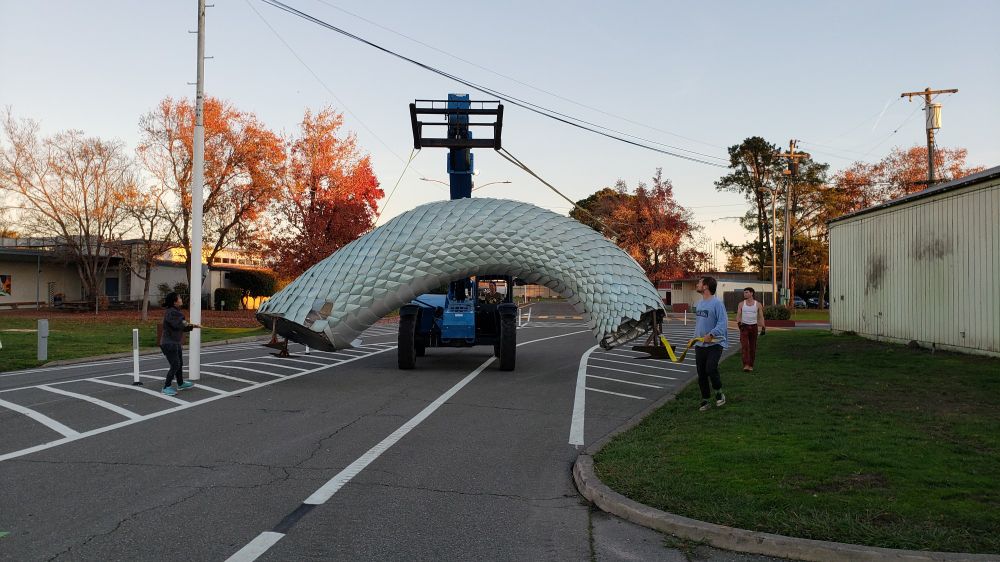 A tiny portion of a very long sea serpent is moved down a road by a forklift. A number of people are pulling lines to help maneuver the piece around a power pole and under some power lines.