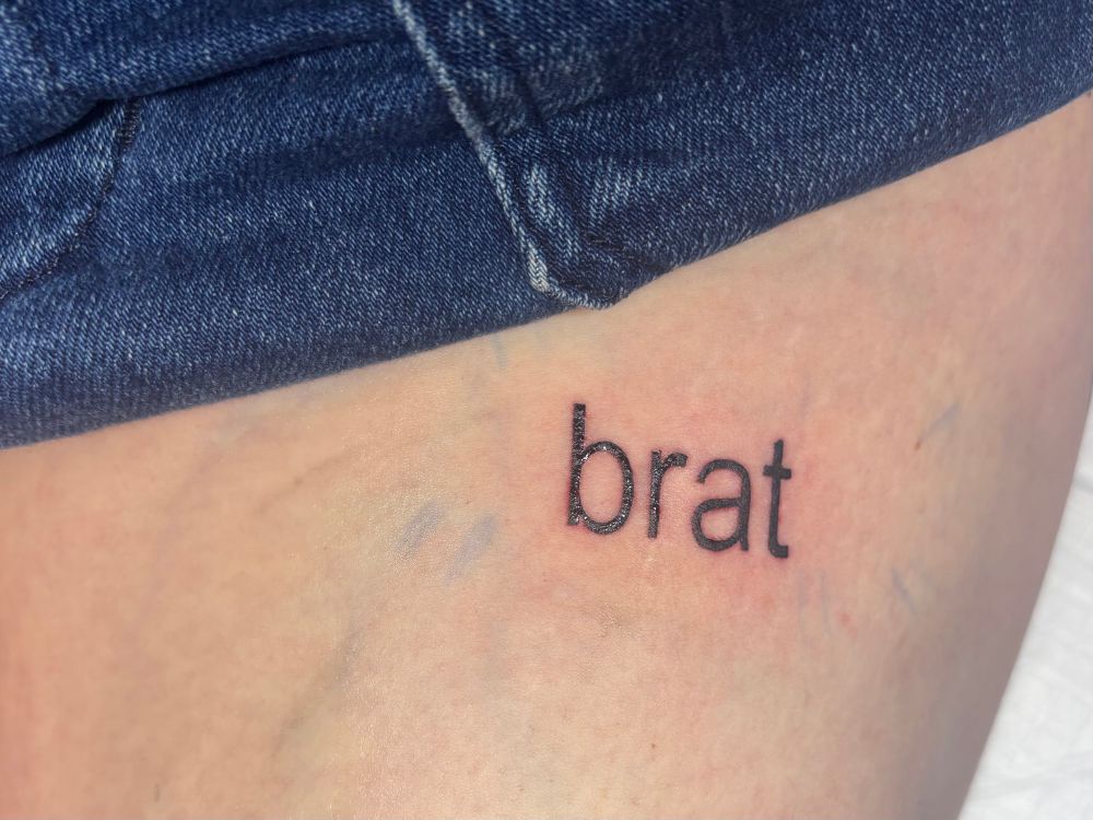 Close up of fresh tattoo on skin of “brat” in Arial font like the Carli XCX album