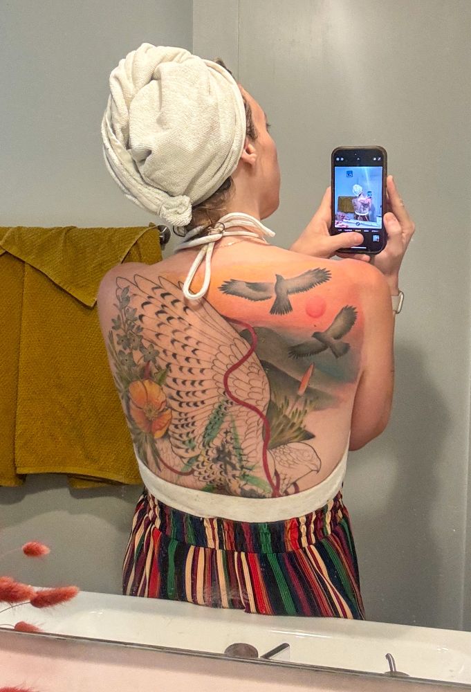 Woman taking selfie of back in mirror. You can see a partially finished coverup full back piece with an outline of a red tailed haw while 2 fly overhead with a sunset over foothills and poppies