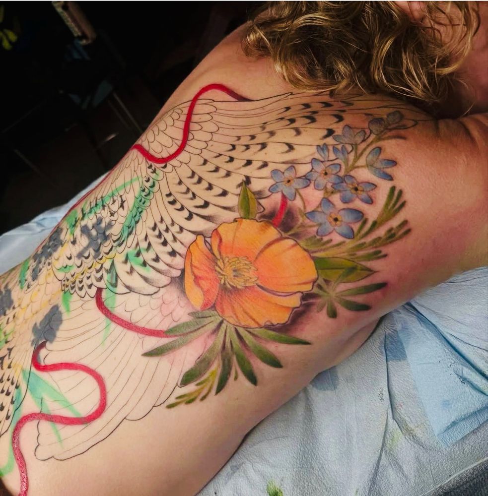 Woman lying face down with partially complete back tattoo of a red tailed hawk and poppy and forget me not, only the flowers are filled in with colo