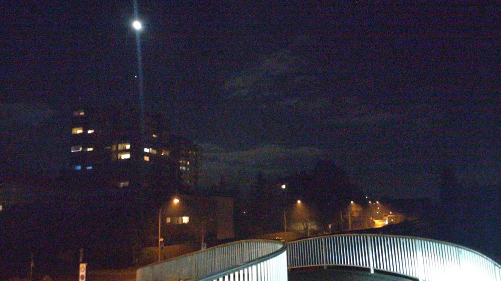 Blick entlang einer gebogenen Fußgänger- und Radfahrerbrücke bei Nacht. Die Metallgeländer sind vom Fahrradscheinwerfer beleuchtet. Im Hintergrund eine mit vermutlich Natriumdampflampen gelb beleuchtete Wohnstraße und ein ca. 7-stöckiger Wohnblock bei dem Fenster in den oberen 4 Etagen beleuchtet sind. Darüber der Mond mit ein bläulichen Lensflare.