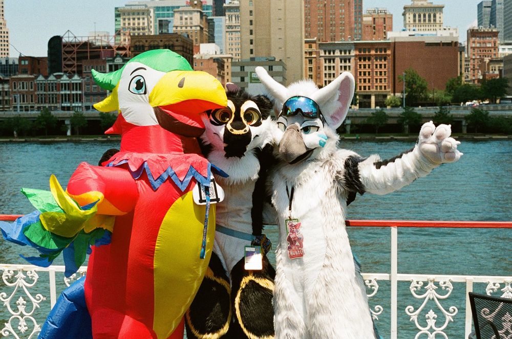 Three fursuiters on a river cruise with a view of the city behind them