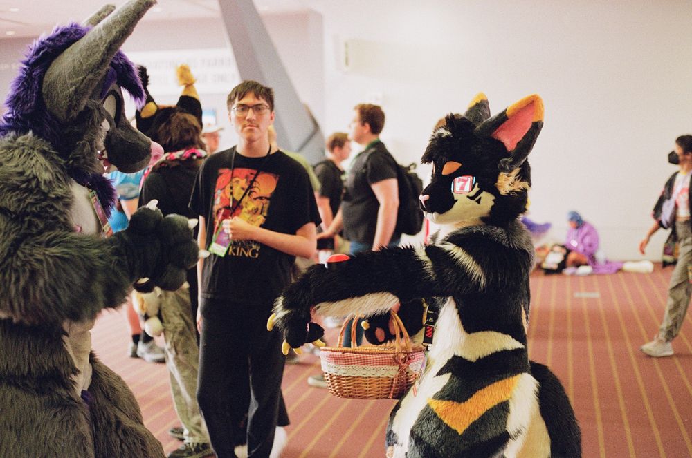 A cat fursuiter with slot machine eyes extends his paw to another fursuiter. There is a big red button on his arm