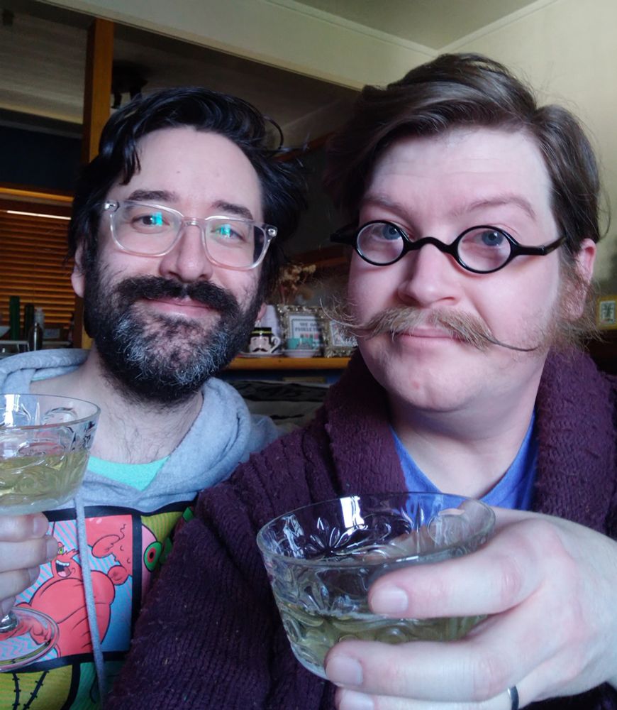 Two men holding champagne glasses, one with a beard, the other a moustache.