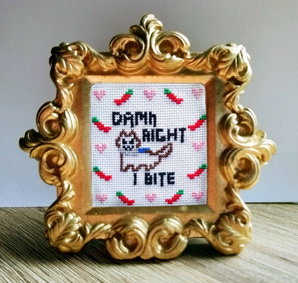 Fancy gold frame with a small cross stitched, angry kitty, with a knife in its mouth, the text says "damn right I bite" - this is frames with little red chilis and pink hearts