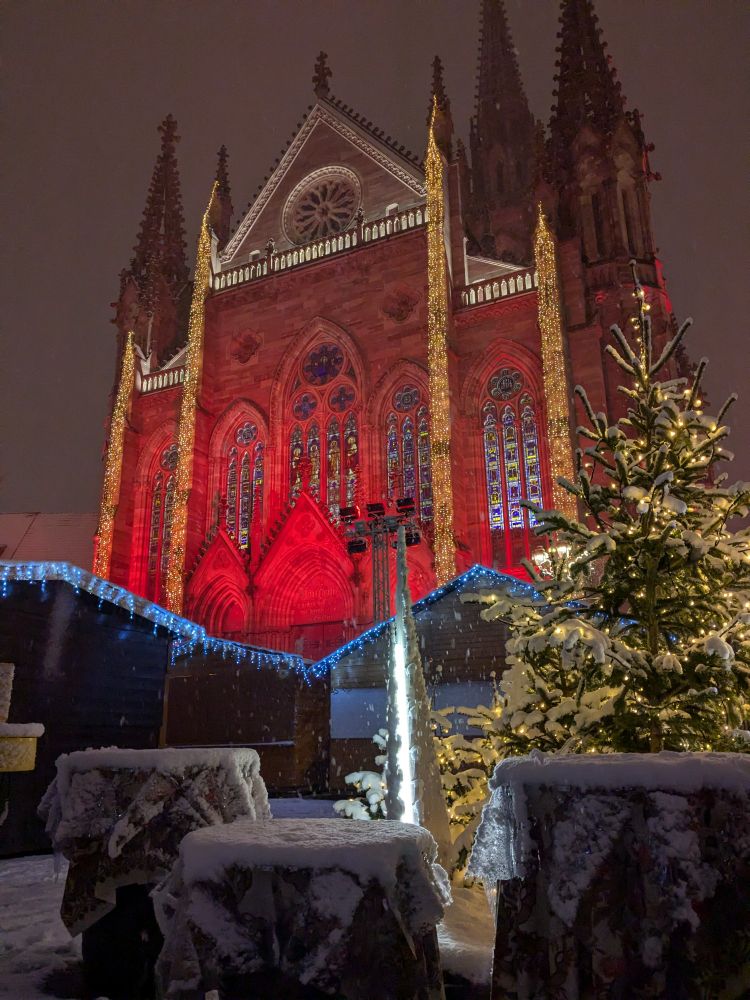 La façade du temple éclairée de rouge et avec des guirlandes verticales. Devant des chalets, un sapin et des tables recouvertes de neige 