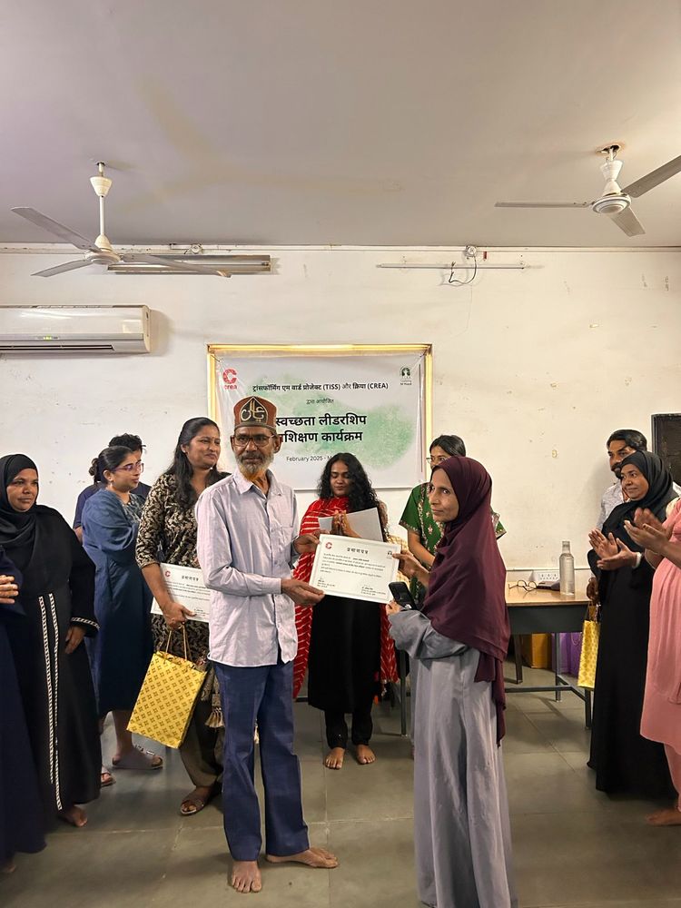 Two people holding their completion certificate while being surrounded by participants and staff members. There's a poster in the background with CREA and TISS logos.

