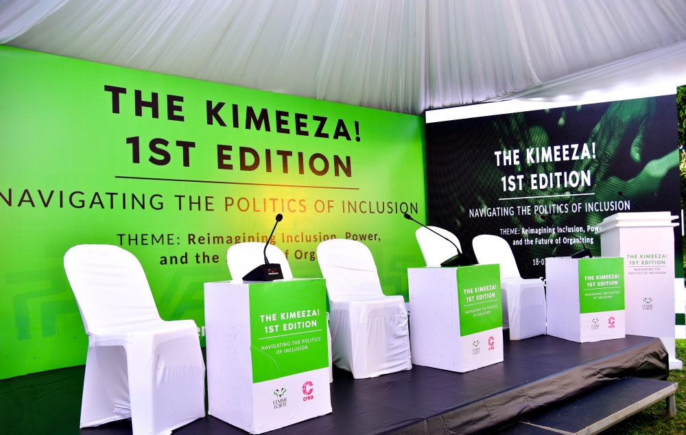 A stage setup from The Kimeeza 1st Edition, co-created by CREA and Femme Forte, featuring white chairs and green podiums under a white tent. The backdrop displays the event’s theme: “Navigating the Politics of Inclusion – Reimagining Inclusion, Power, and the Future of Organising.”