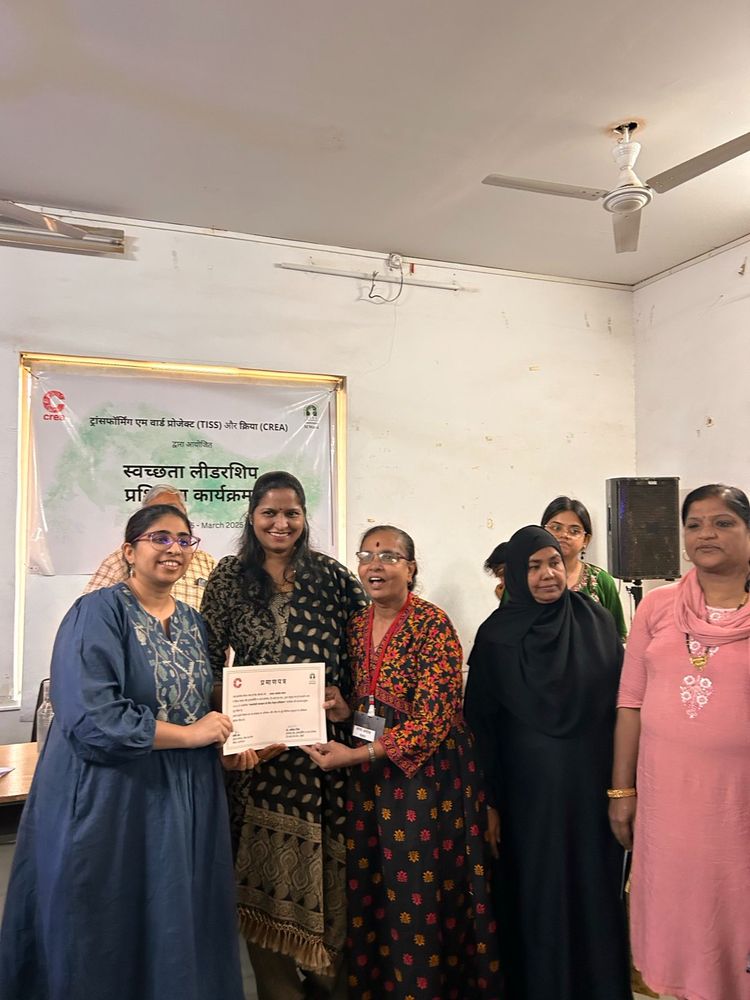Three people holding the certificate, posing for the camera while being surrounded by other participants. There's a poster in the background with CREA and TISS logos.

